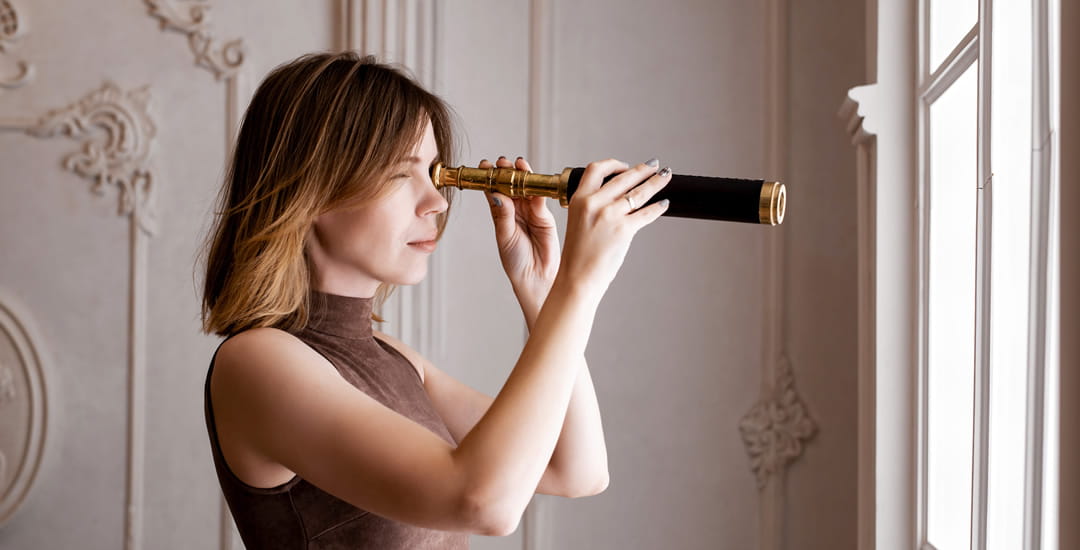 Woman looking out of the window at neighbours’ home with telescope