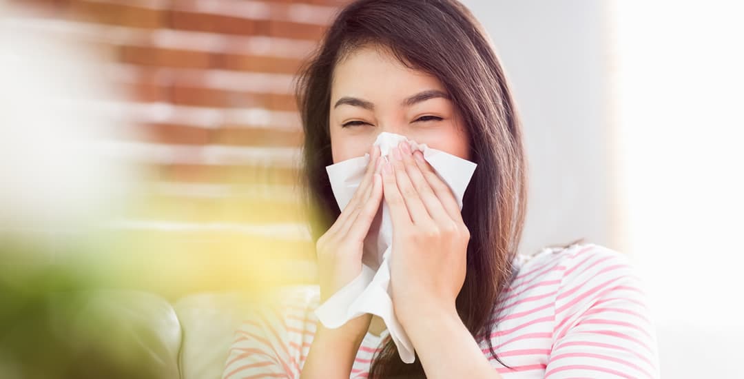 Woman sneezing into tissue in front of window blinds