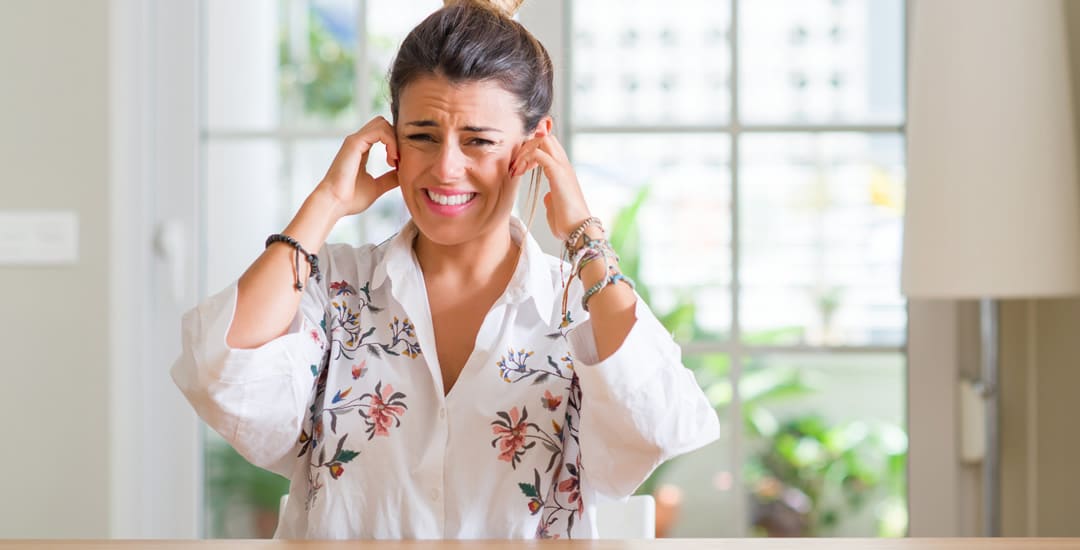 Woman at kitchen window covering ears with fingers due to noise